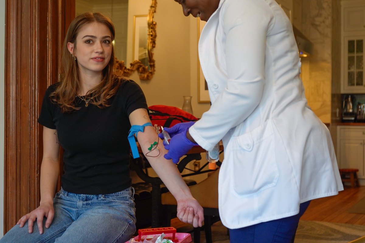 A young woman sits for a blood draw while a clinician in a white coat works at her arm.