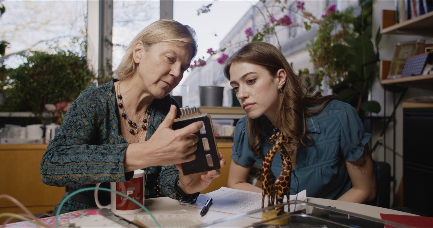 Dr. Linda Griffith shows a small platform device to the director across a workbench in a plant-filled office.