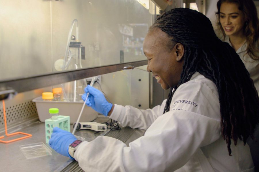 A researcher in a lab coat pipetting at a bench, laughing, with a colleague behind her.