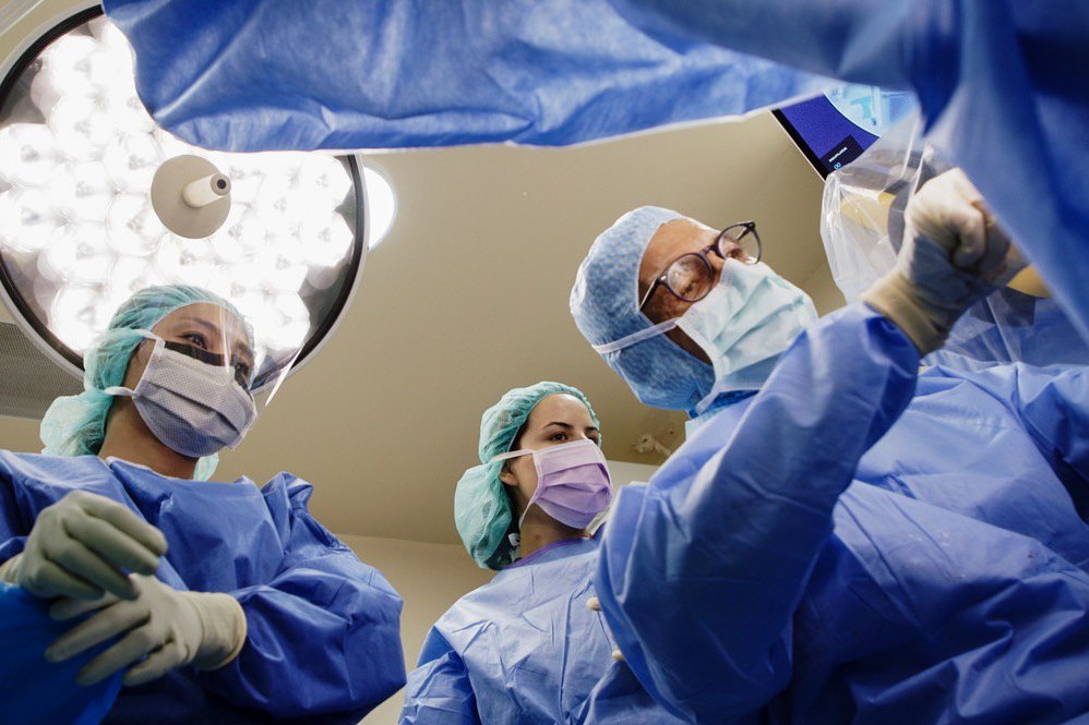An operating room mid-procedure, seen past a masked nurse in the foreground.