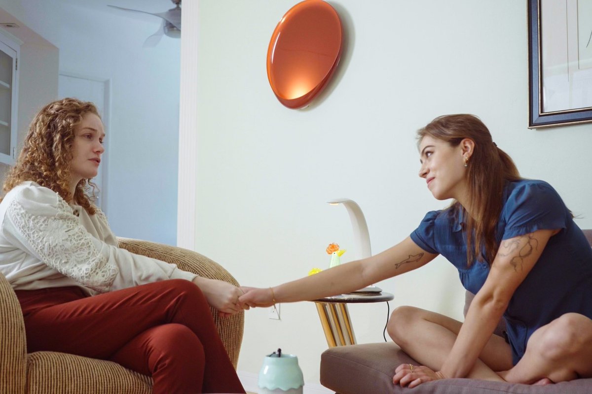 Two women sitting across from each other in a living room, reaching to hold hands.