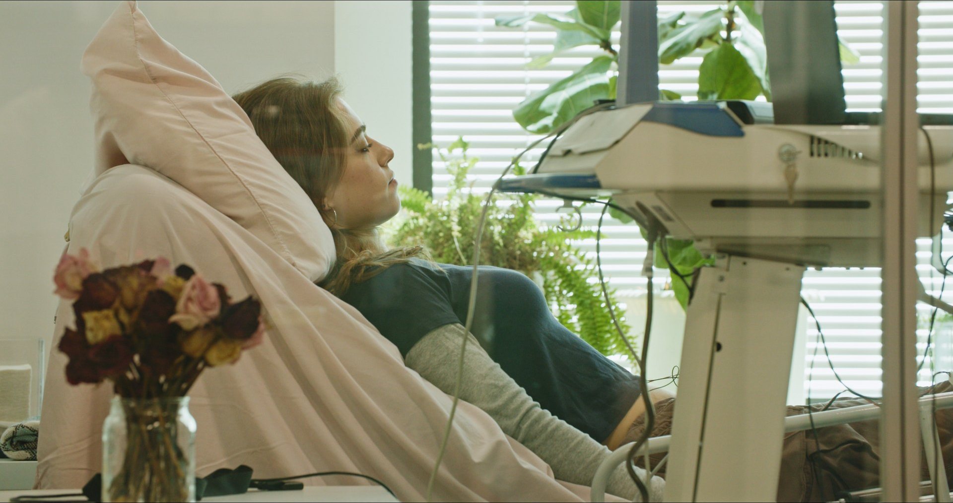 A patient reclining in a treatment chair beside monitoring equipment, flowers and plants nearby.