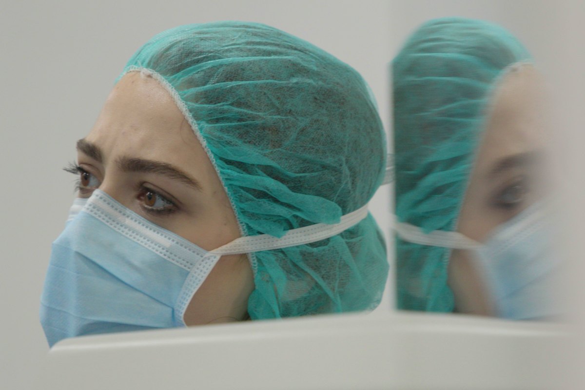The director in surgical cap and mask, seen in profile and reflected in the glass beside her.
