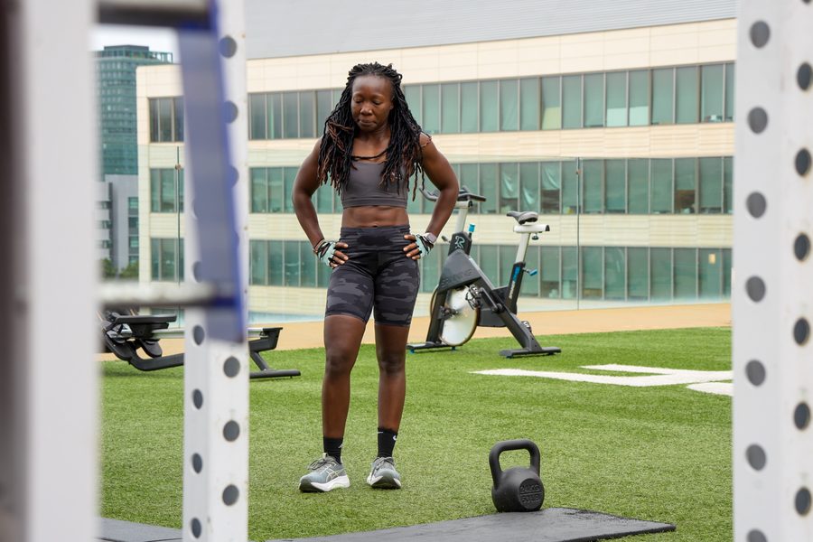 A woman in training gear standing with hands on hips on a rooftop gym deck, city buildings behind her.